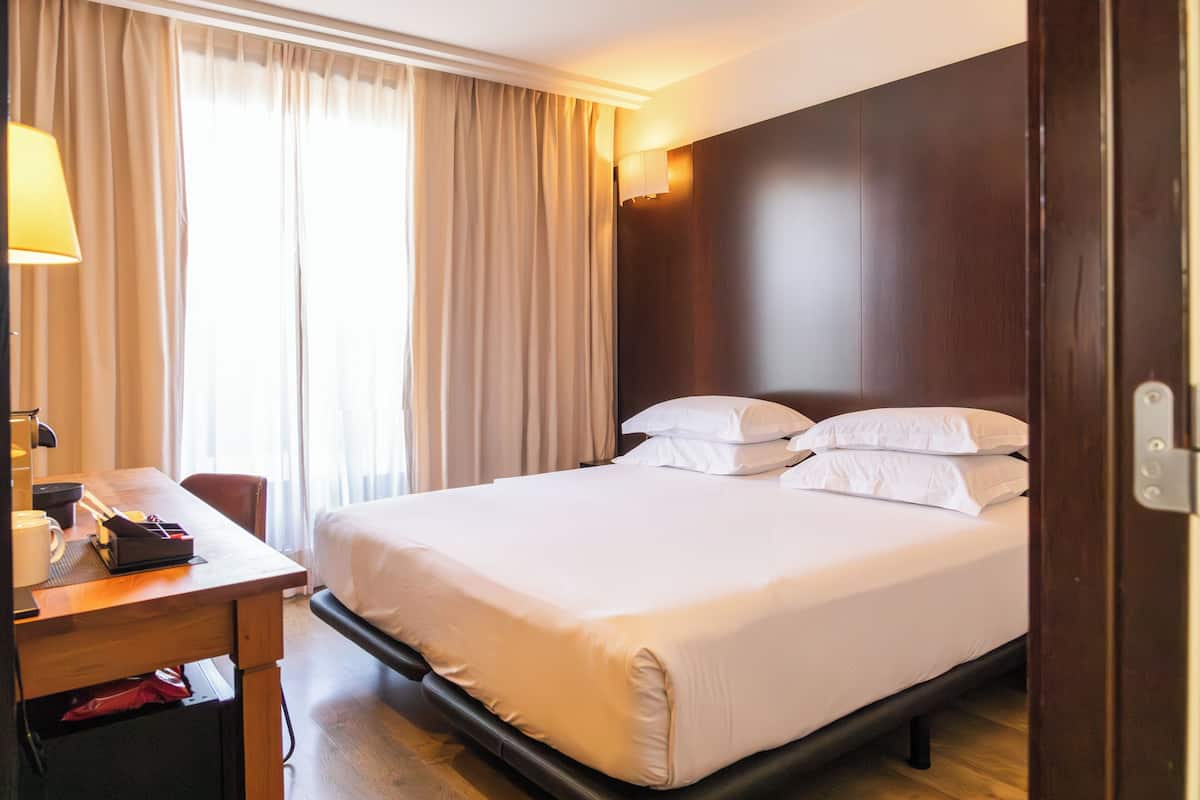 A hotel room with a bed, a desk, a lamp, and a window with curtains.