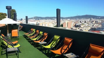 A rooftop terrace with rows of colorful lounge chairs and a table, overlooking a cityscape with buildings and hills.
