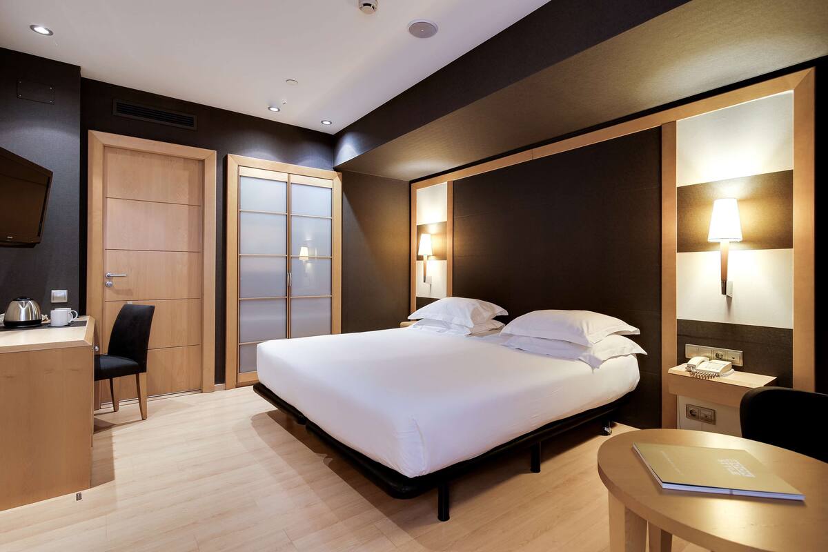 A hotel room with a bed, bedside table, lamp, desk, chair, and a small table with a notebook.