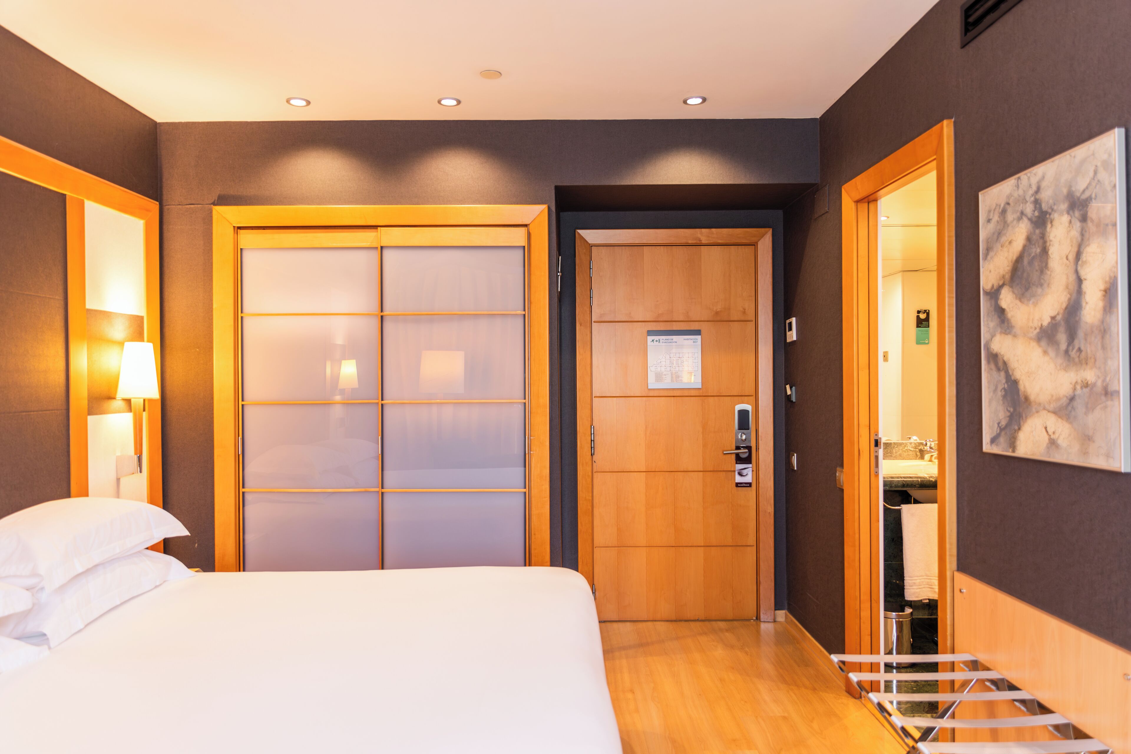 Habitación de hotel con cama, armario de madera, espejo, toallero y un cuadro en la pared.