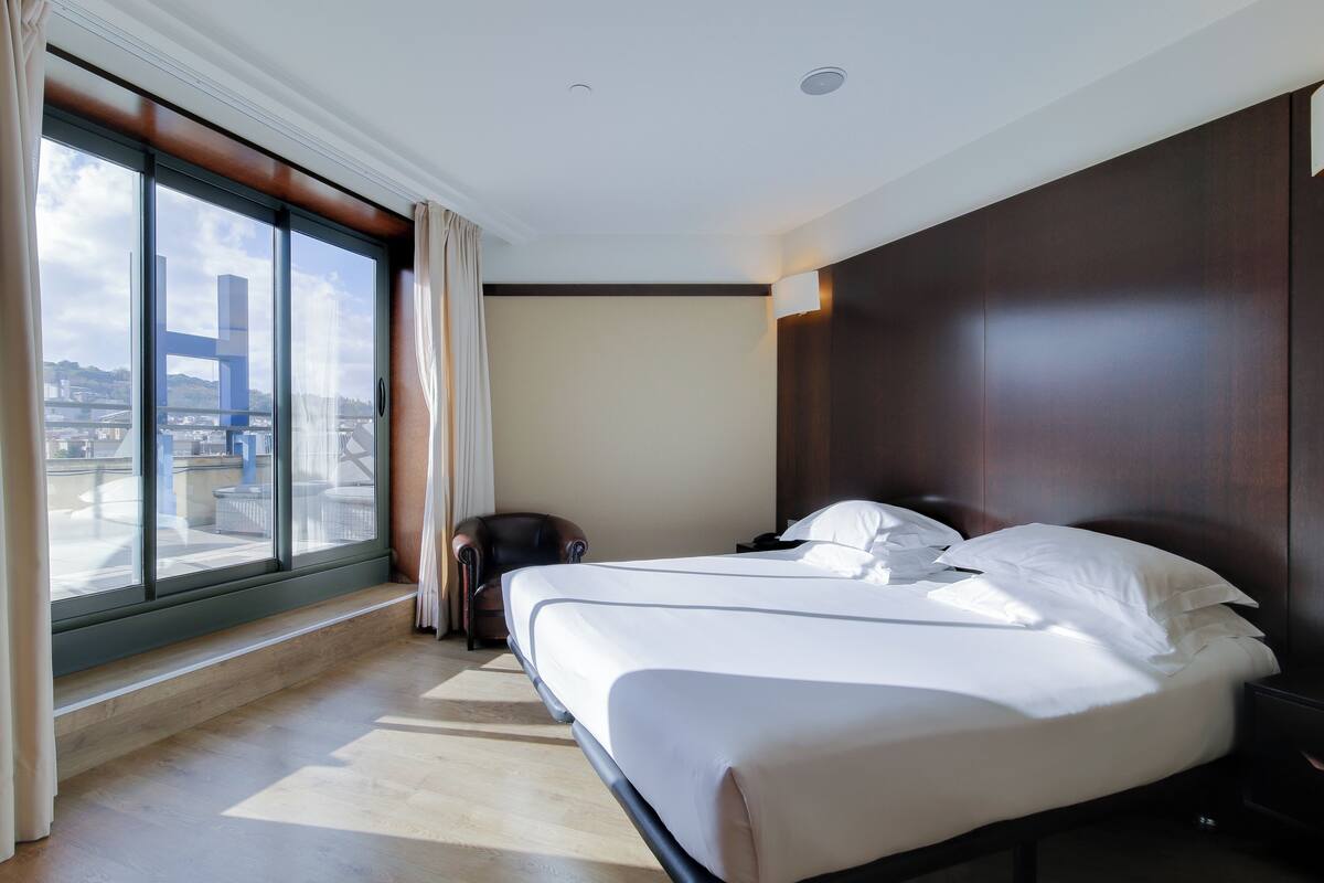 A hotel room with a large bed, a chair, a nightstand, and a window with a view of a bridge.