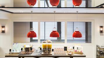 Montaje de desayuno buffet con diversas opciones de comida y bebida, entre ellas jugos y pasteles, en un interior moderno con ventanas amplias y lámparas colgantes rojas.