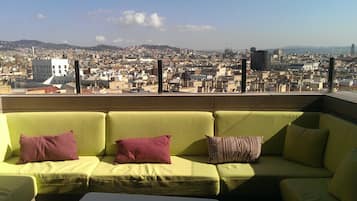 A rooftop terrace with a green sofa and cushions, overlooking a cityscape with buildings and hills.