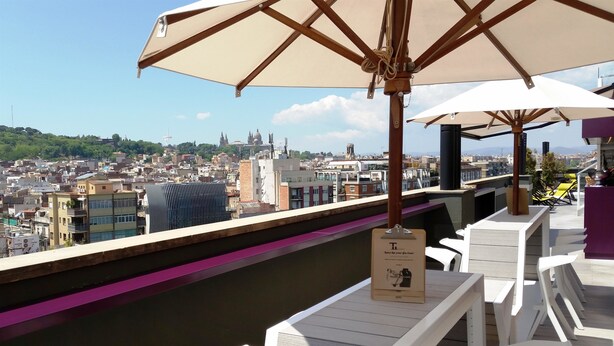 A rooftop bar with white furniture and a large umbrella, overlooking a cityscape with buildings and a river. - Hotel Barcelona Universal (Barcelona)