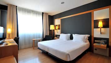Una habitación de hotel con una cama grande, un escritorio con lámpara, una silla, una mesita y un televisor.