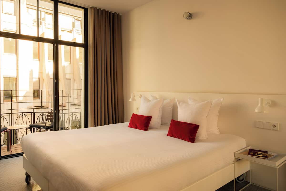 A hotel room with a large bed, a nightstand, a window with curtains, and a view of the outside.
