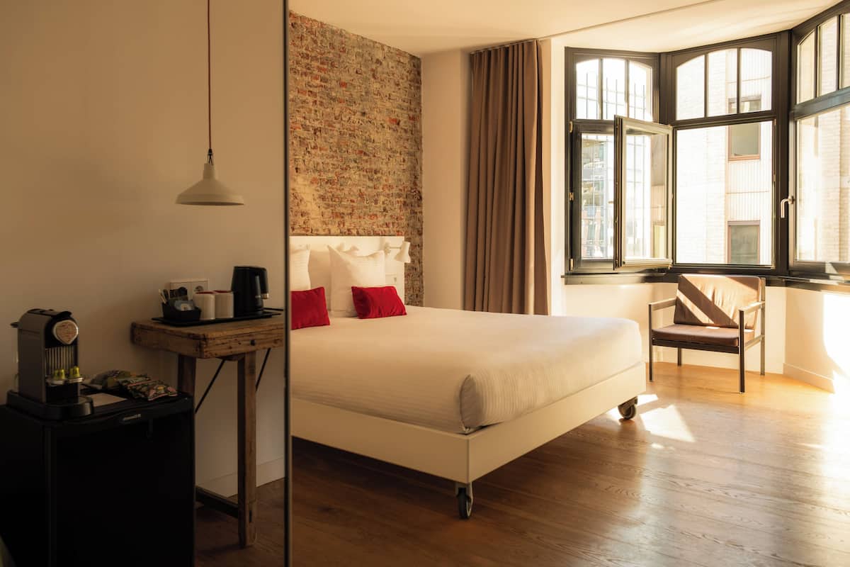 A hotel room with a bed, a desk with a coffee maker, a chair, and a window with curtains.