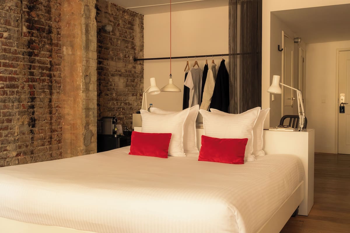 A hotel room with a brick wall, a bed with white bedding and red pillows, a hanging light fixture, and a coat rack with clothes.