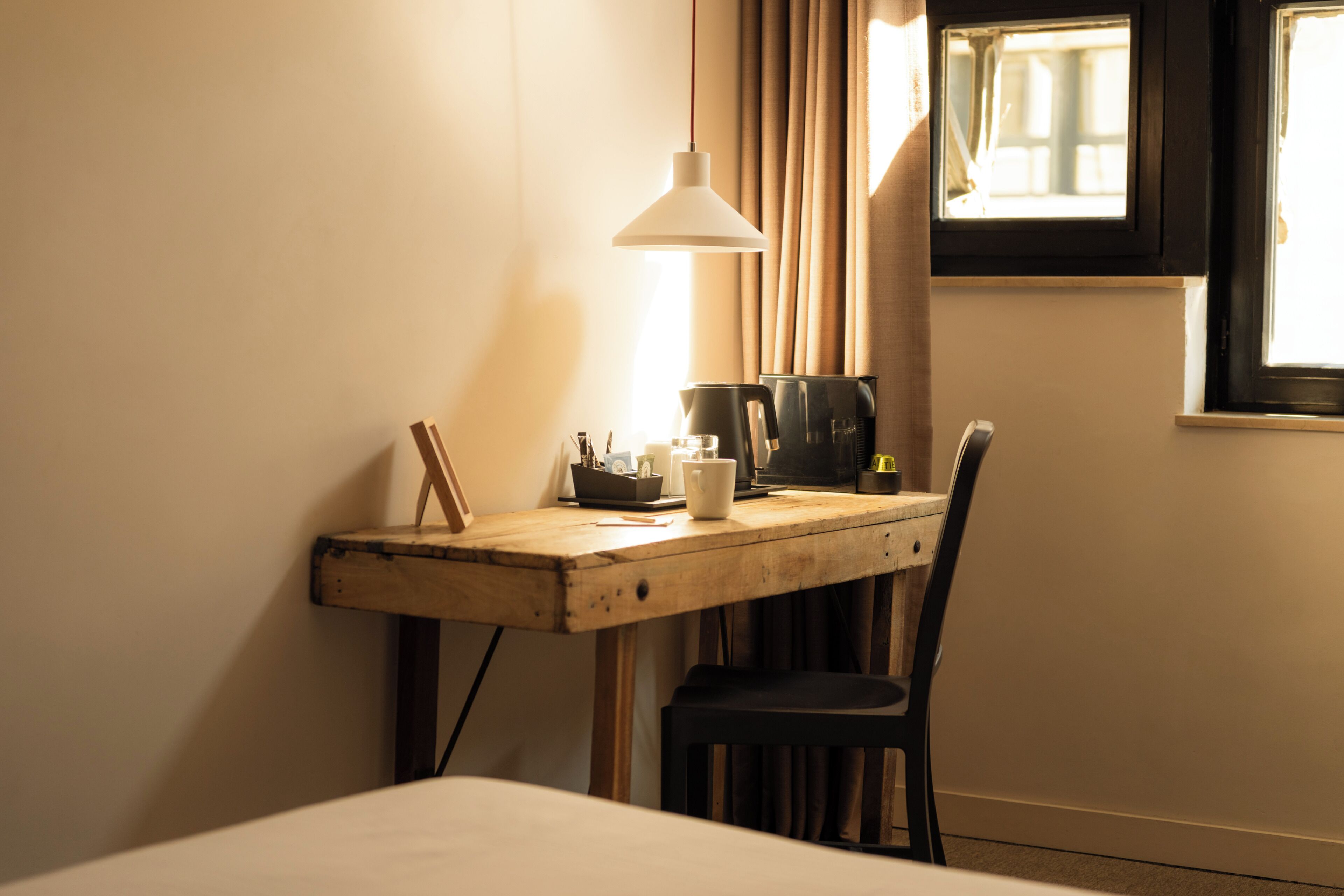 A wooden desk with a lamp, a chair, a coffee maker, and a window with curtains.