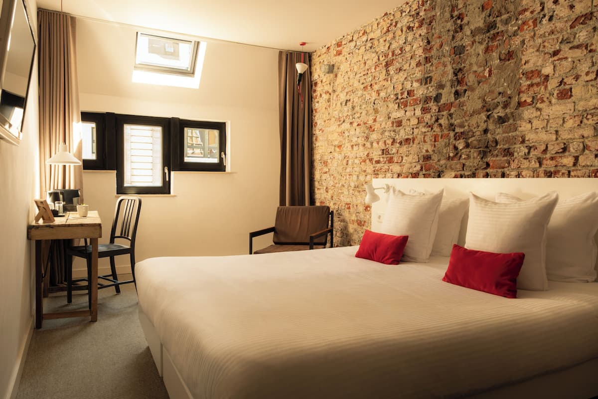 A hotel room with a large bed, a desk with a chair, a small table, and a brick-patterned wall.
