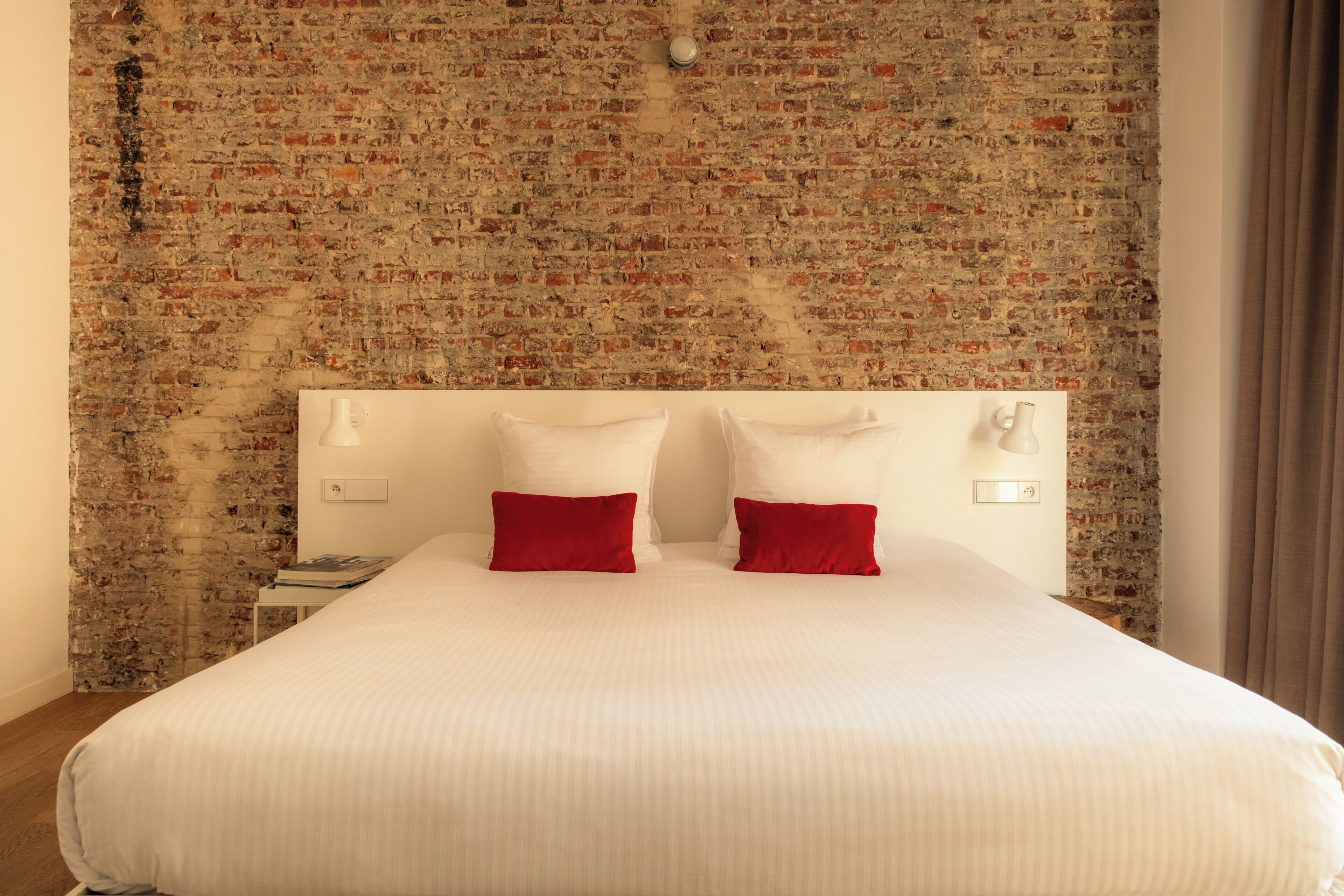 A hotel room with a brick-patterned wall, a large bed with white bedding and red pillows, a nightstand, and a wall-mounted light fixture.