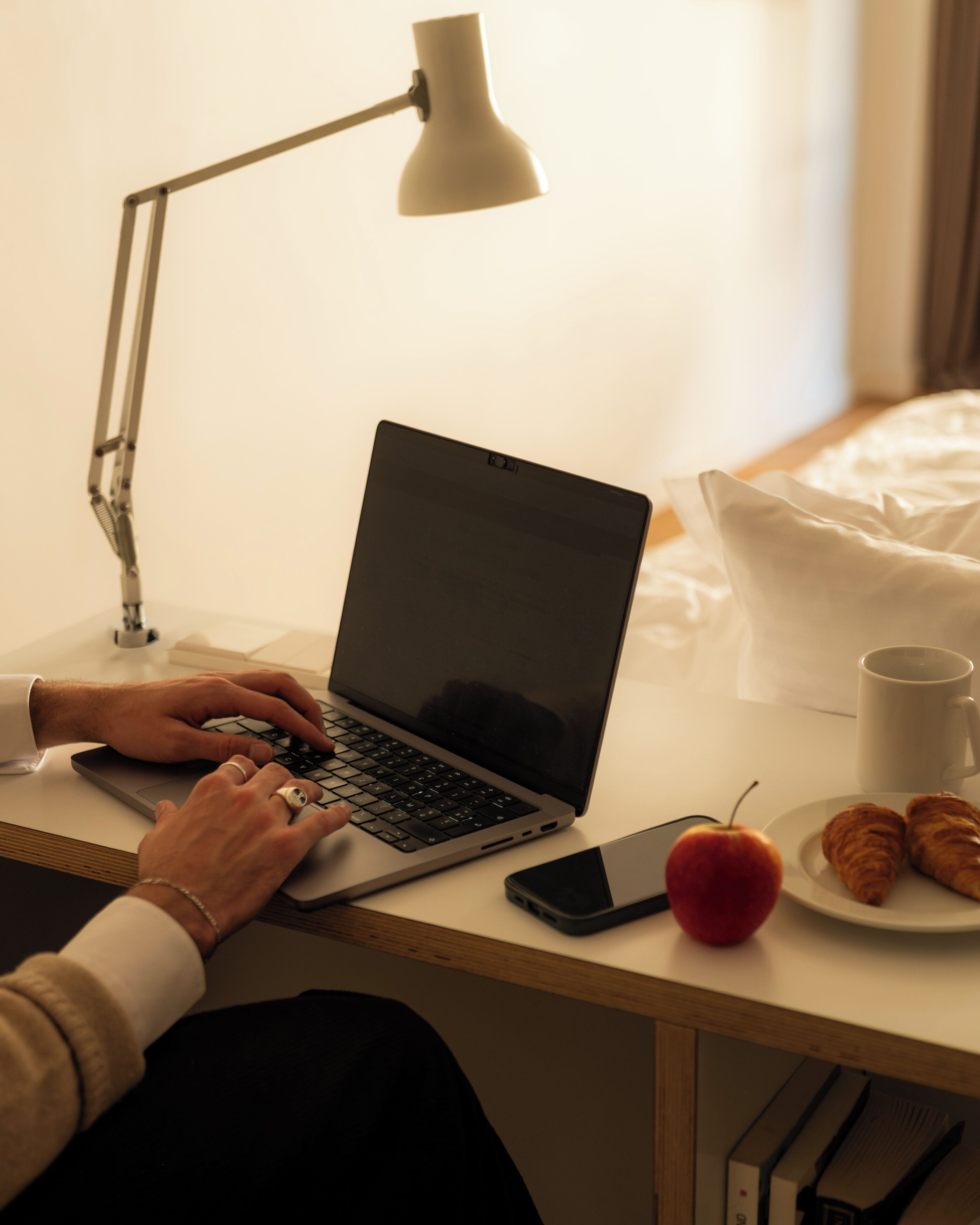 Un portatile su una scrivania con una lampada, uno smartphone, una mela e croissant.