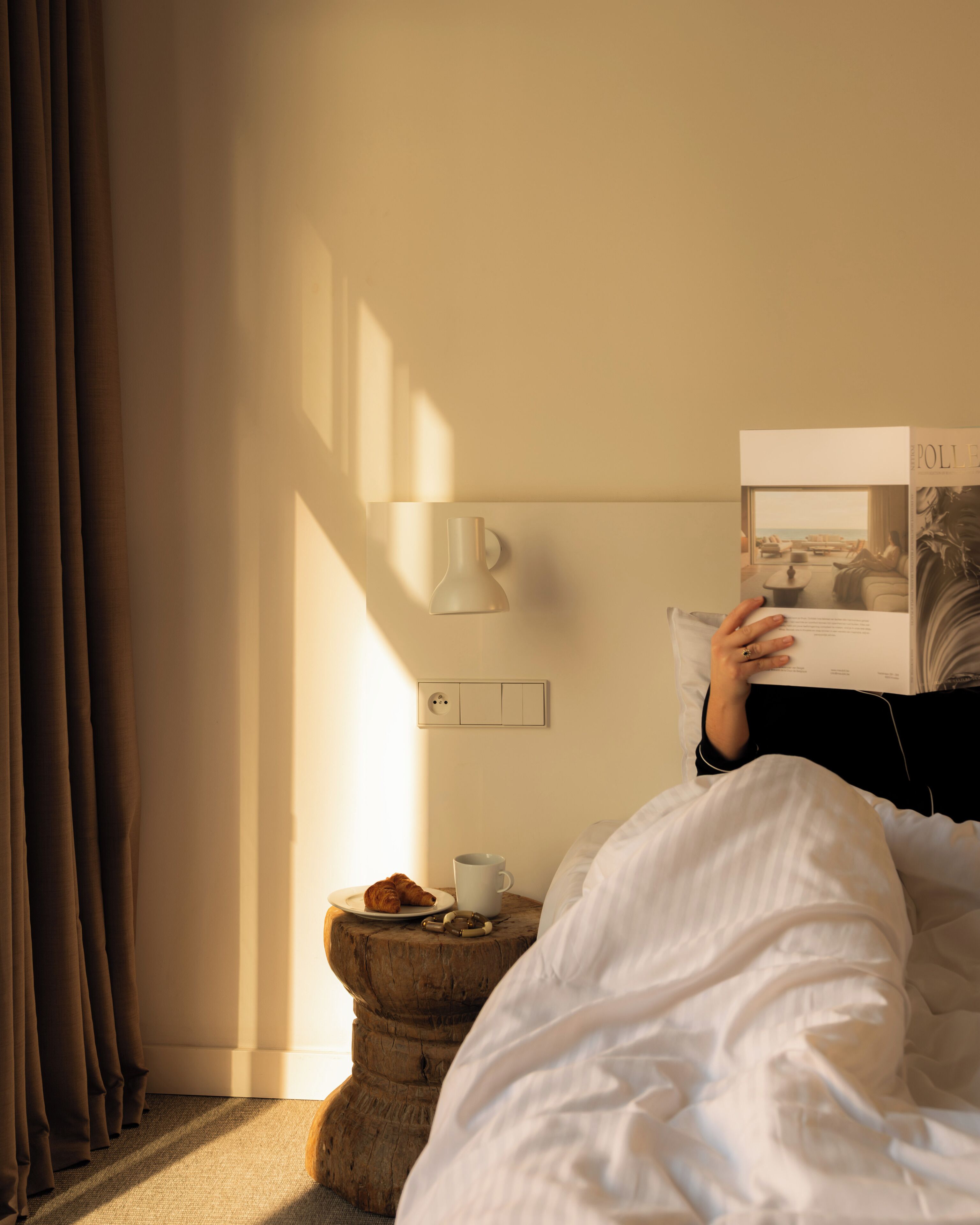 Une personne est assise dans son lit, un livre à la main, et peut admirer le paysage par la fenêtre. Sur une table de chevet, on aperçoit une tasse et un croissant. Une lampe est fixée au mur.