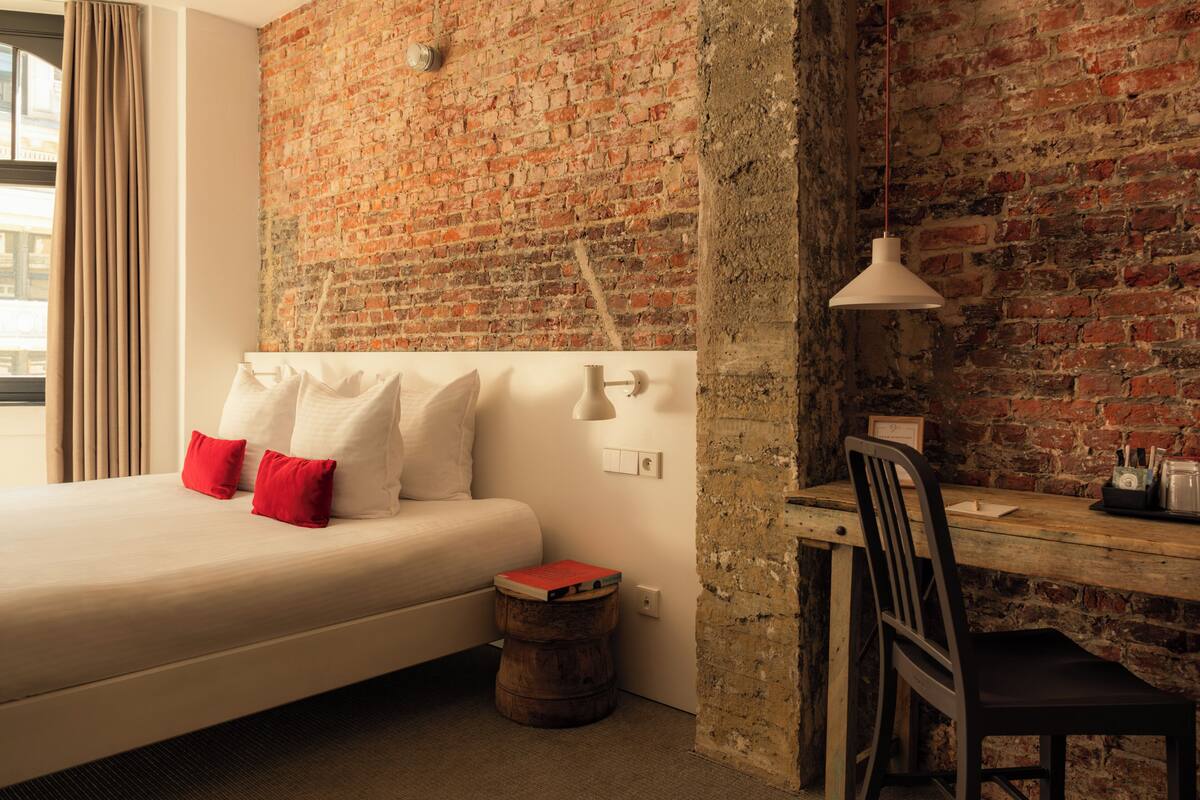 A hotel room with a brick wall, a bed with white and red pillows, a desk with a lamp, and a small wooden stool.