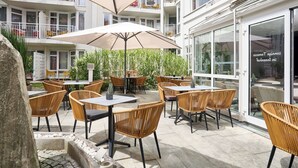 Un área de patio al aire libre con sillas y mesas de mimbre, un paraguas grande y vistas a un edificio con balcones.