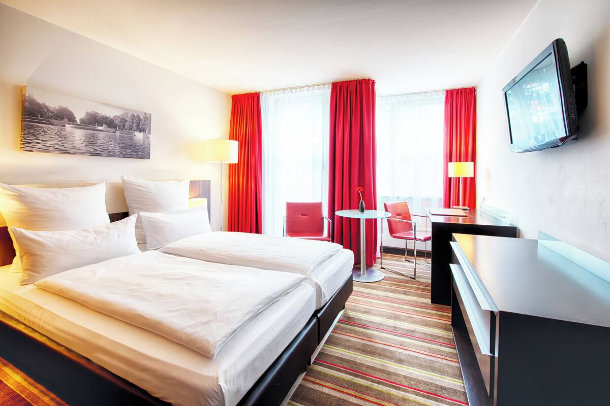 Een hotelkamer met twee bedden, een bureau, een stoel, een televisie en een raam met rode gordijnen.