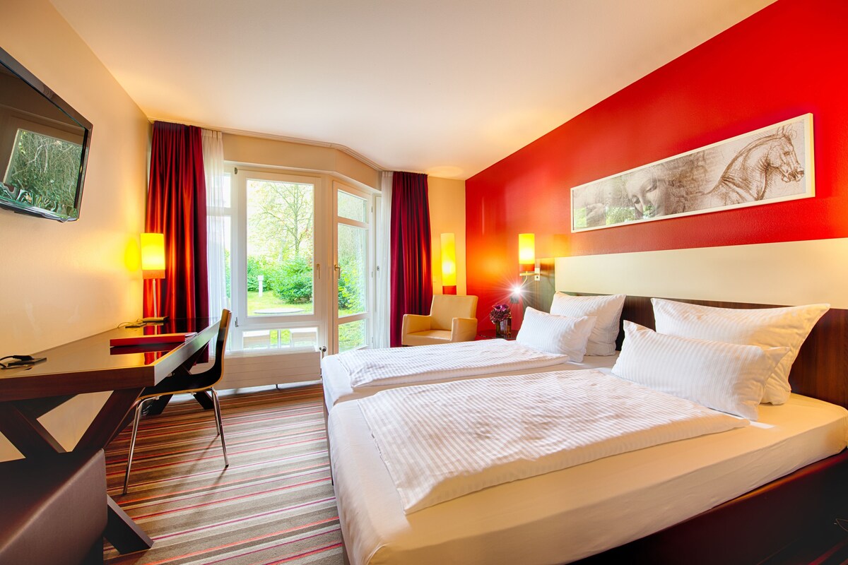 Ein Hotelzimmer mit einem Bett, zwei Nachttischen, einem Schreibtisch, einem Sessel und einem großen Fenster.