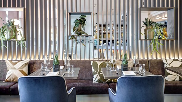 Un comedor moderno con sofá, dos mesas y sillas, que incluye un panel de madera vertical y plantas colgantes.