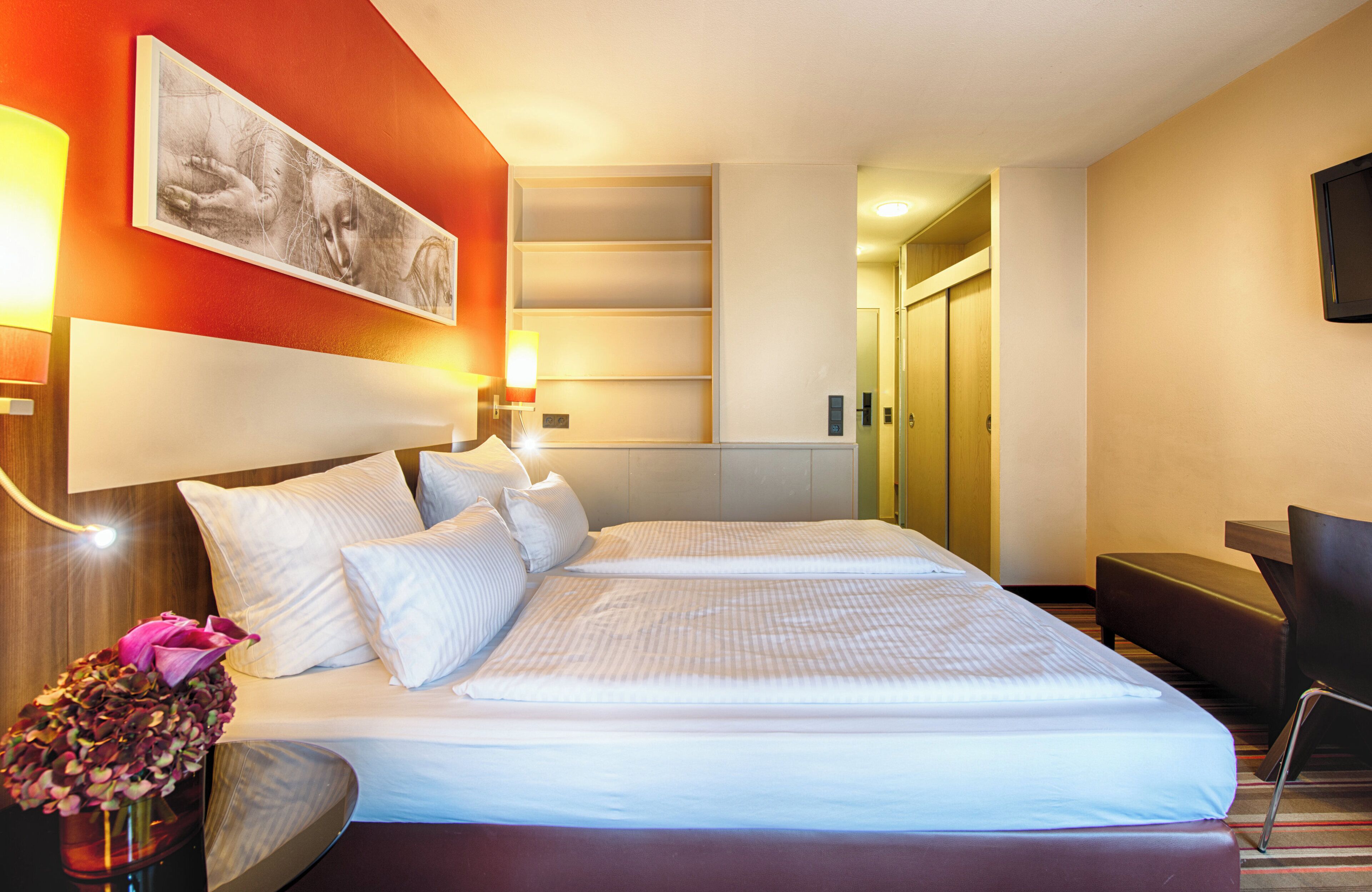 Habitación de hotel con cama, lámparas de noche, escritorio, silla, un jarrón con flores y un cuadro en la pared.