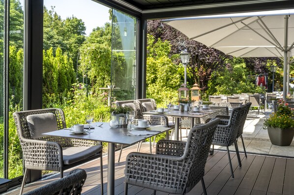 An outdoor dining area with wicker chairs and tables, large windows, and a view of lush greenery. - Best Western Plus Hotel Erb (Vaterstetten)