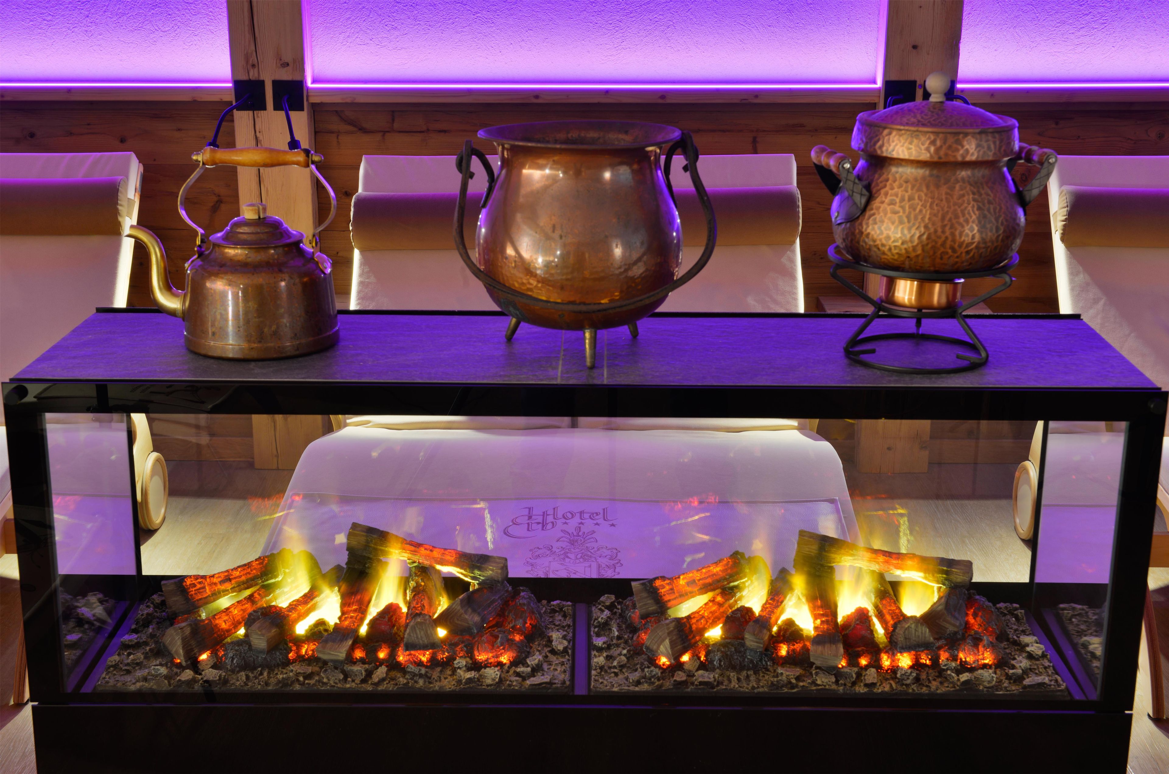 A kitchen area with a wooden table displaying three copper pots and a kettle, and a lower shelf with a glass display of burning wood.