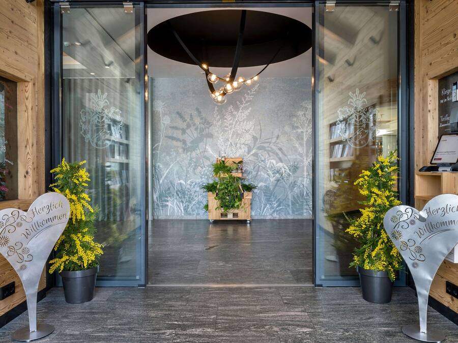 Entrada de hotel con diseño moderno, que incluye una puerta de cristal, un mostrador de recepción de madera y un arreglo decorativo de plantas.