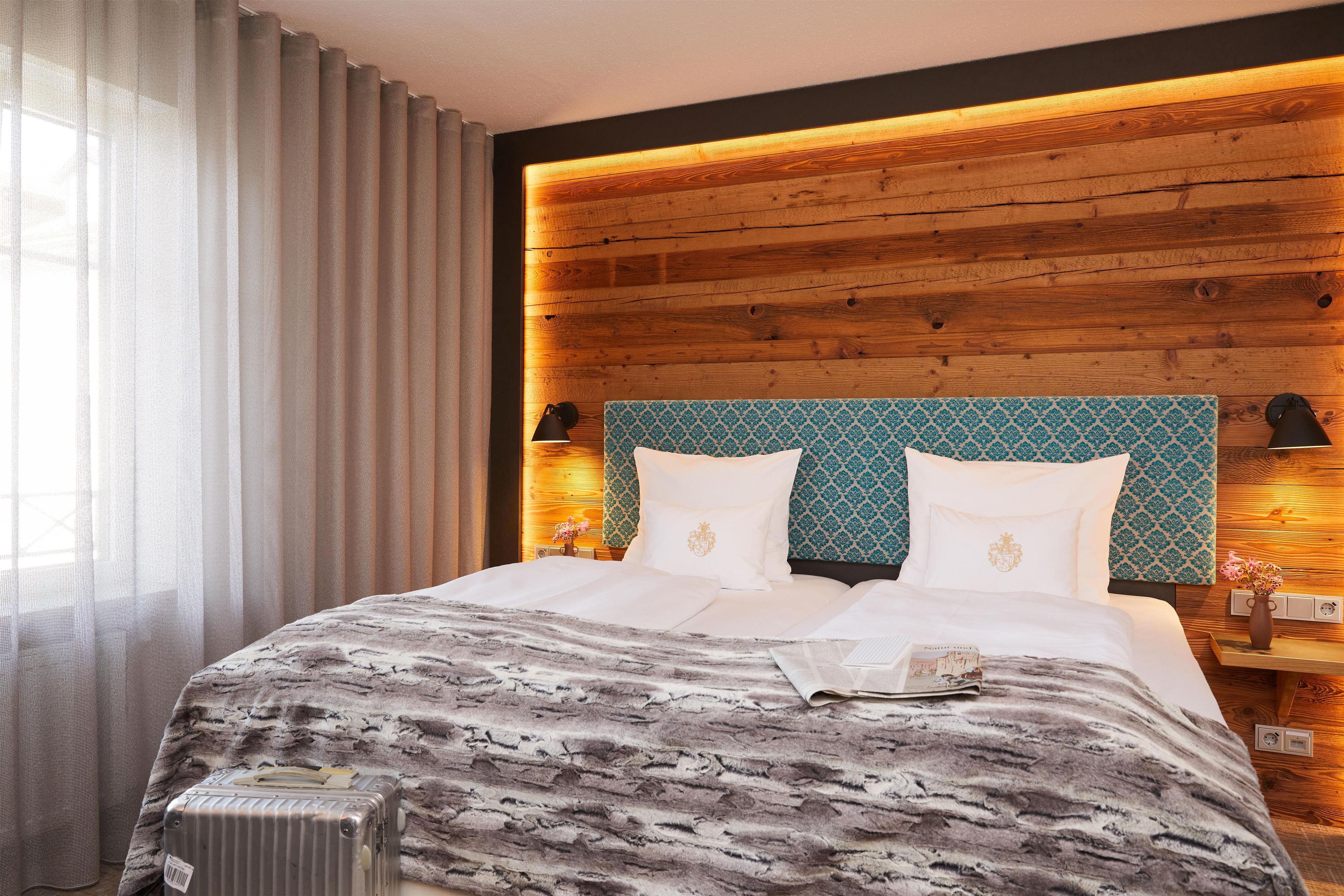 Habitación de hotel con cabecera de madera, cama con ropa blanca, manta decorativa y una maleta en el suelo.