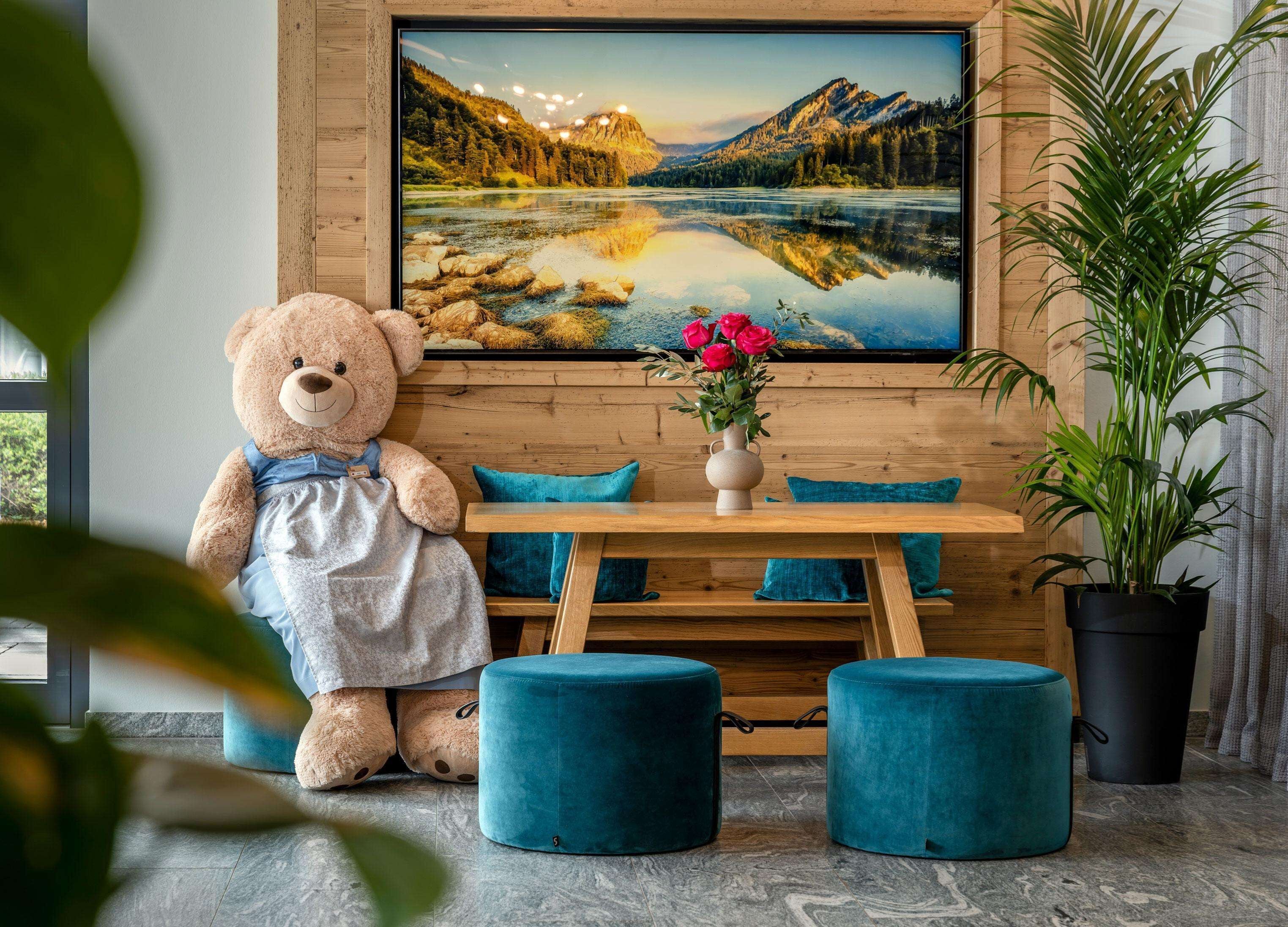 Ein gemütliches Zimmer mit einem großen Teddybären, einem Holztisch mit Blumen in einer Vase, türkisfarbenen Kissen und einem großen Gemälde einer Berglandschaft.