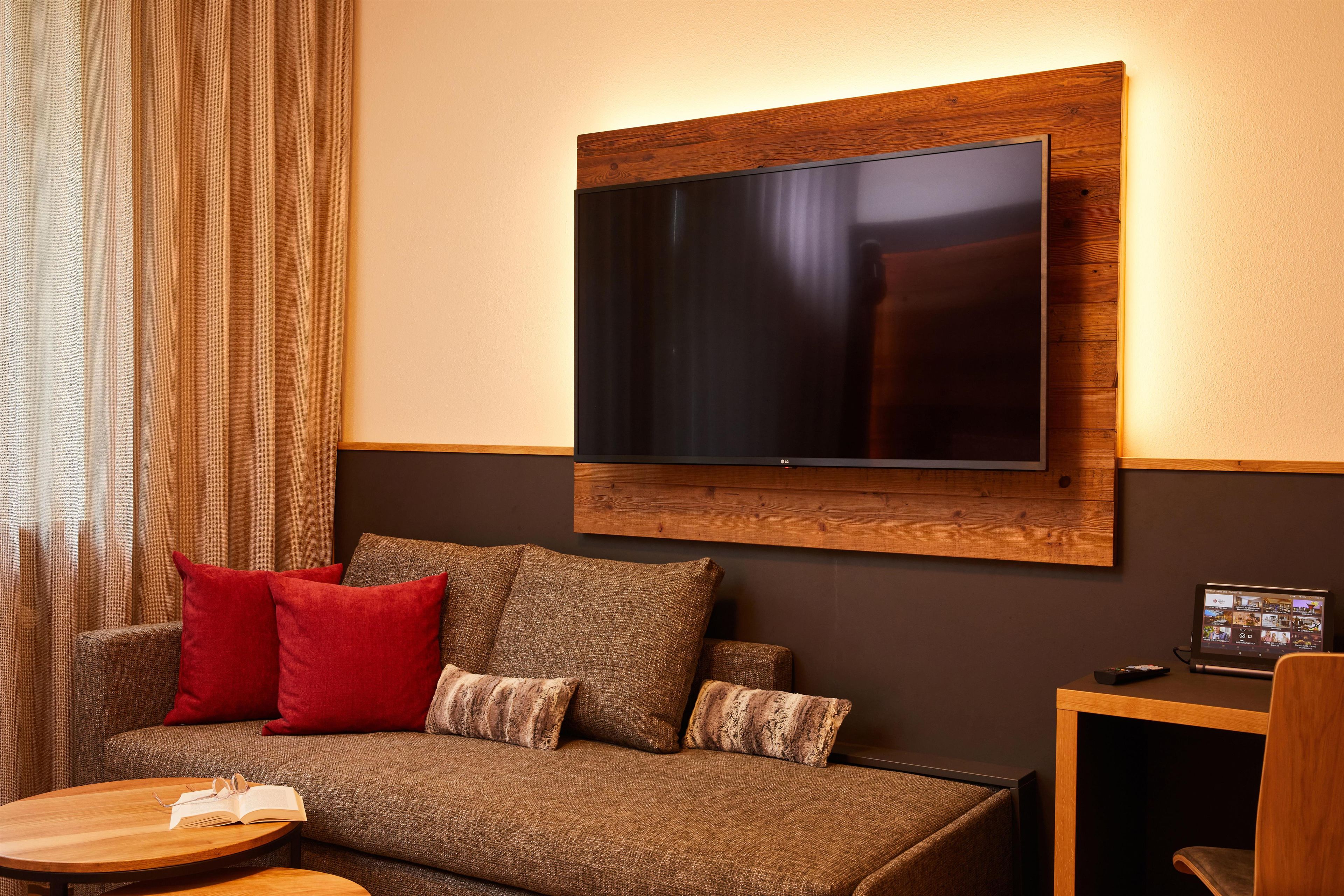 A modern living room with a flat-screen TV mounted on a wooden panel, a brown sofa with red and brown cushions, and a small wooden side table.
