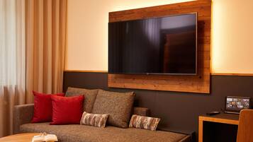 A modern living room with a flat-screen TV mounted on a wooden panel, a brown sofa with red and brown cushions, and a small wooden side table.