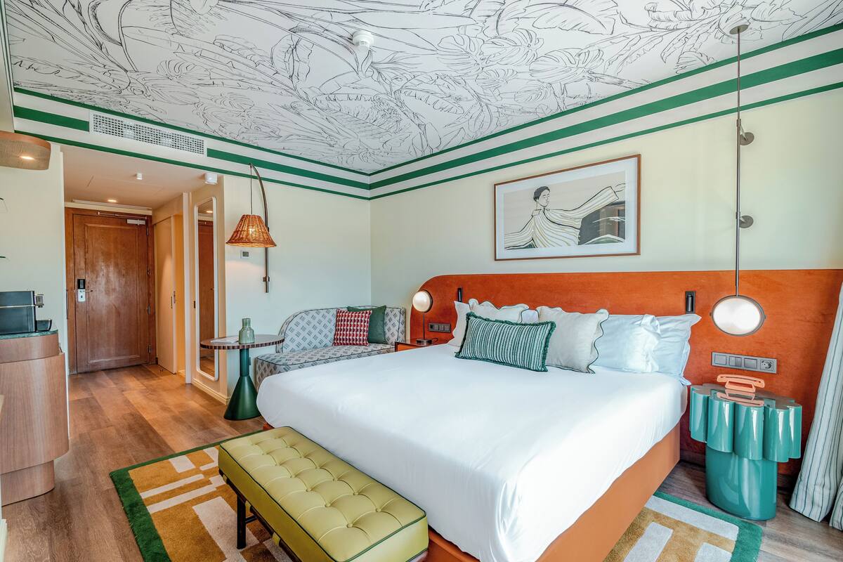 A hotel room with a large bed, a bedside table, a lamp, a bench, and a ceiling with a floral pattern.