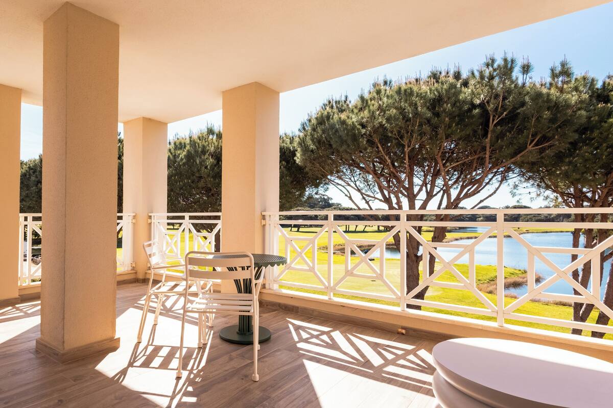 A balcony with a table and chairs, a white railing, and a view of a lake and trees.