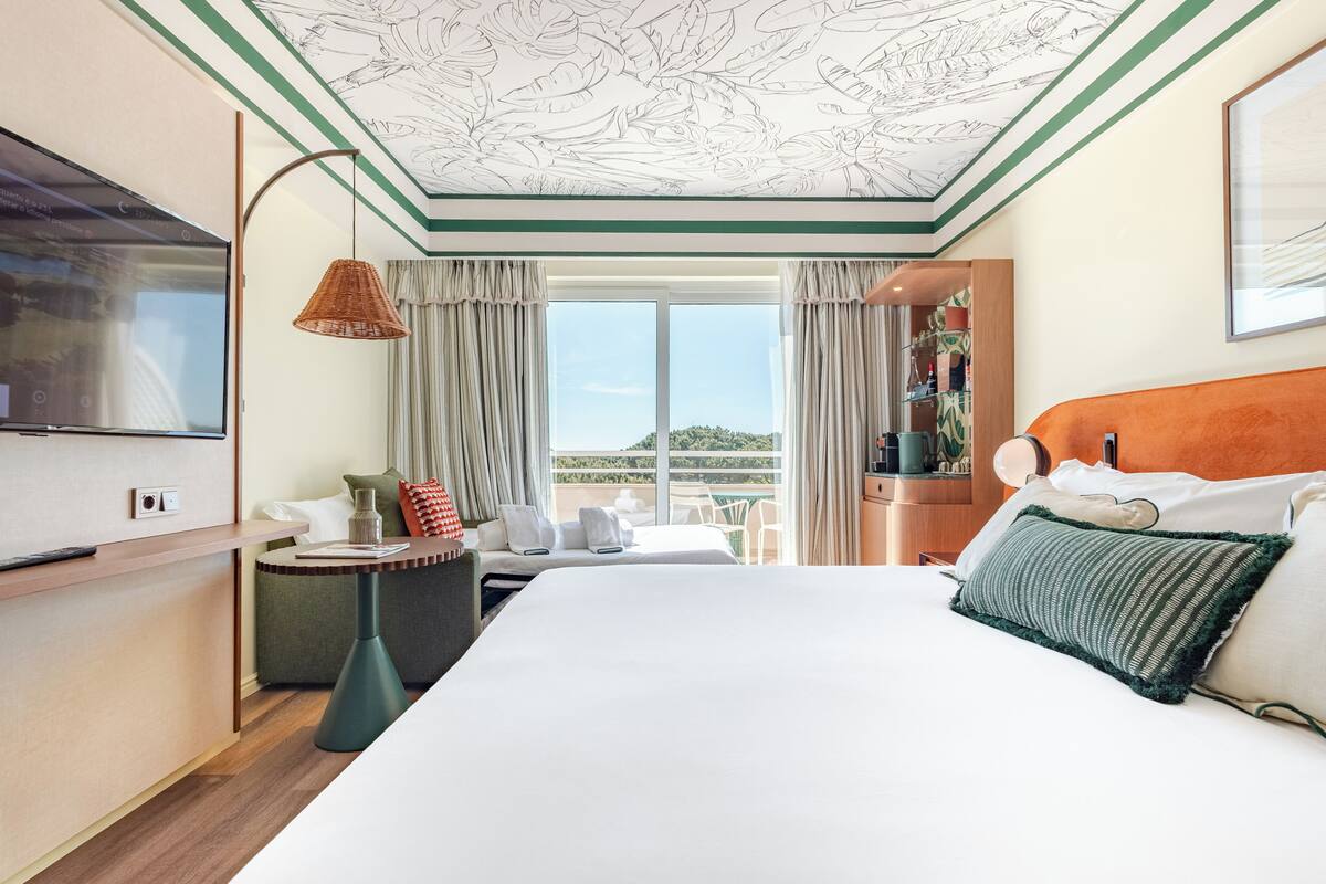 A hotel room with a large bed, a TV, a desk, a chair, and a balcony with a view.
