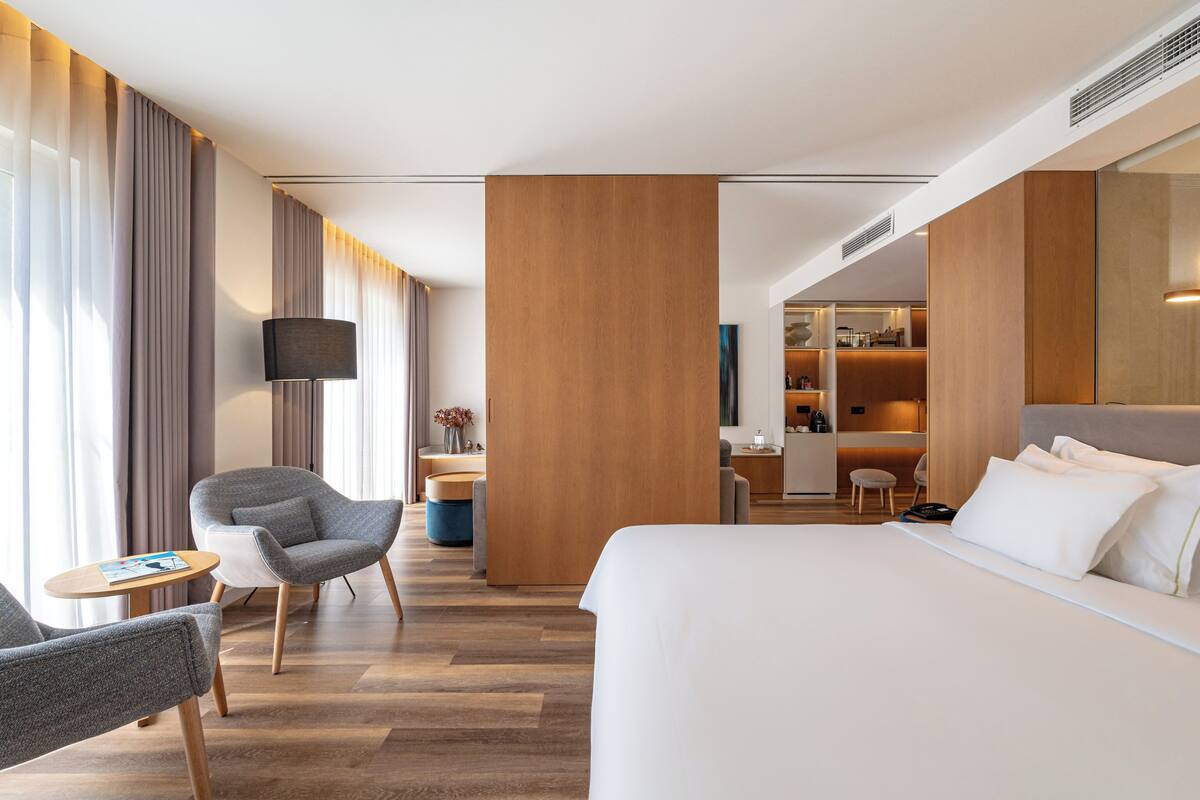 A modern hotel room with a large bed, a seating area with a chair and a small table, a TV, and a kitchenette in the background.