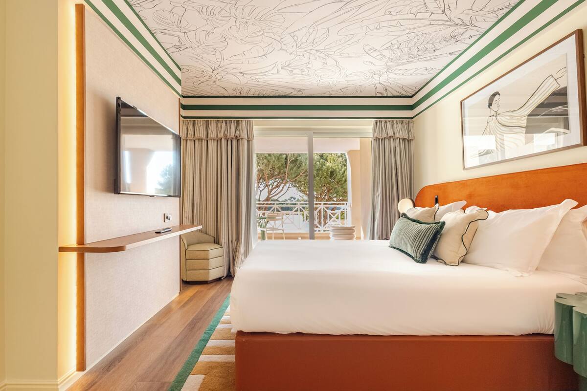 A hotel room with a bed, a TV, a balcony with a view, and a ceiling with a floral pattern.