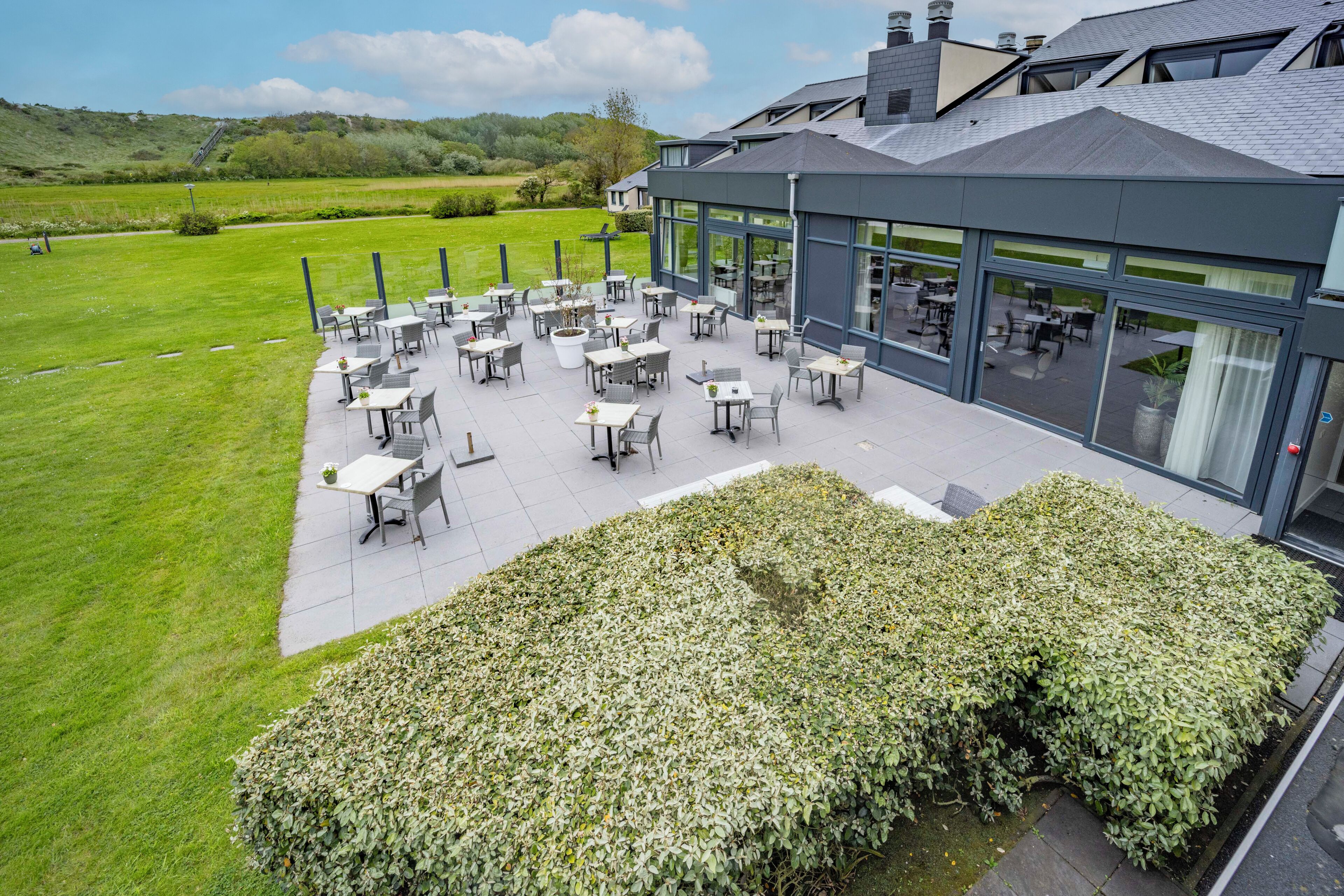An outdoor patio area with tables and chairs, a hedge, and a view of a green field and buildings in the background.