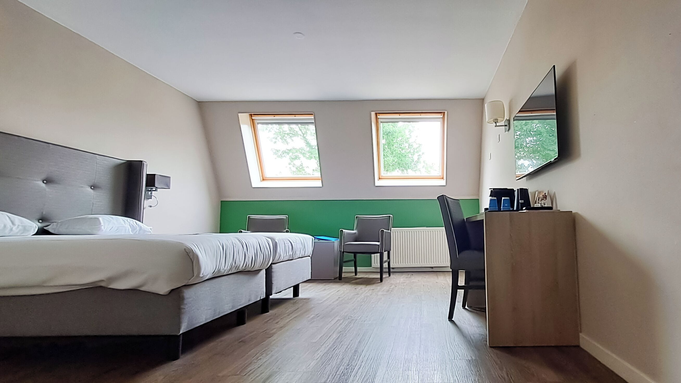 A hotel room with a bed, two windows, a desk, a chair, and a television.