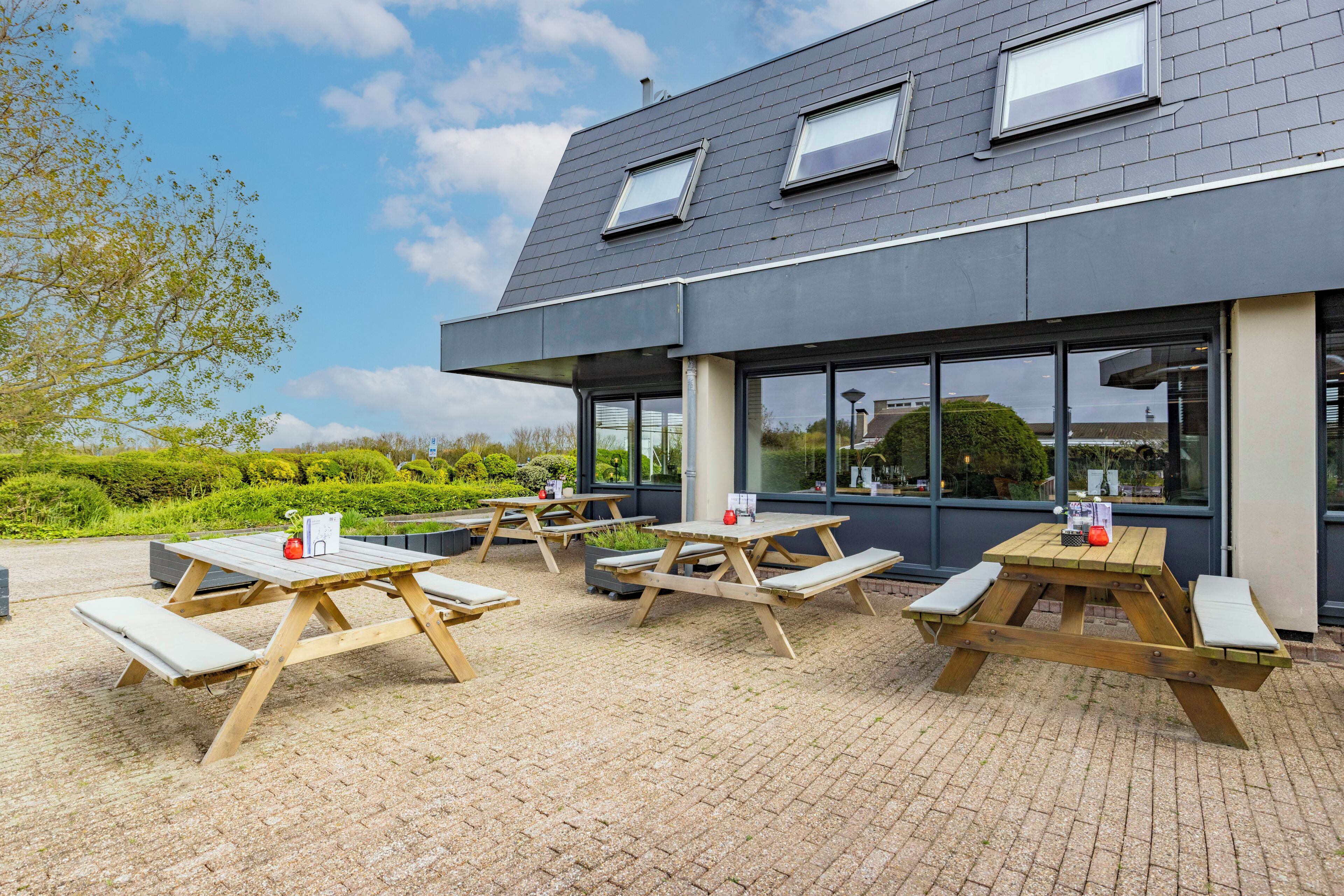 Een terras buiten met houten picknicktafels en banken, een modern gebouw met grote ramen, en een heldere lucht.