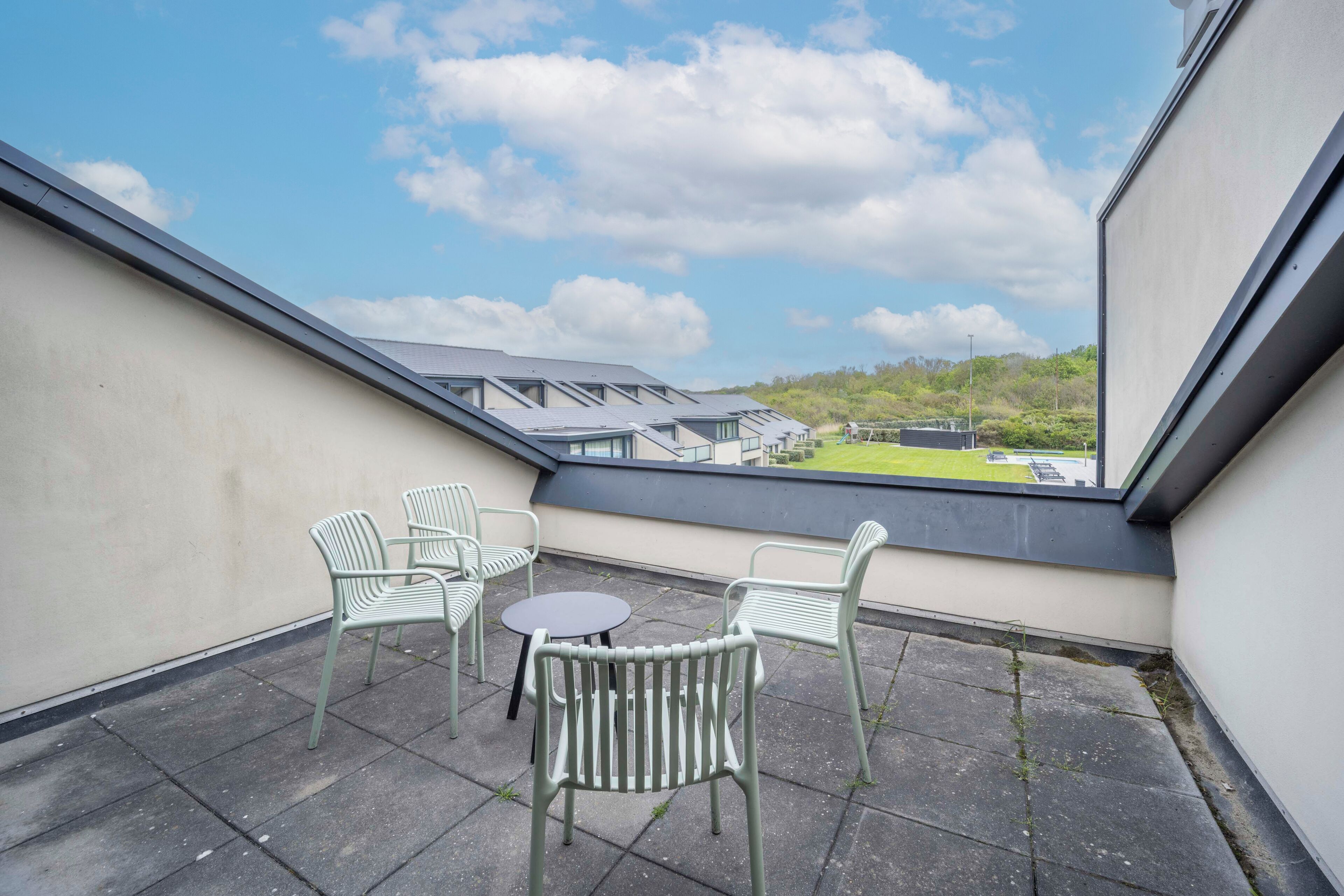 En balkon med et bord og stole, udsigt over et grønt landskab, og en bygning i baggrunden.