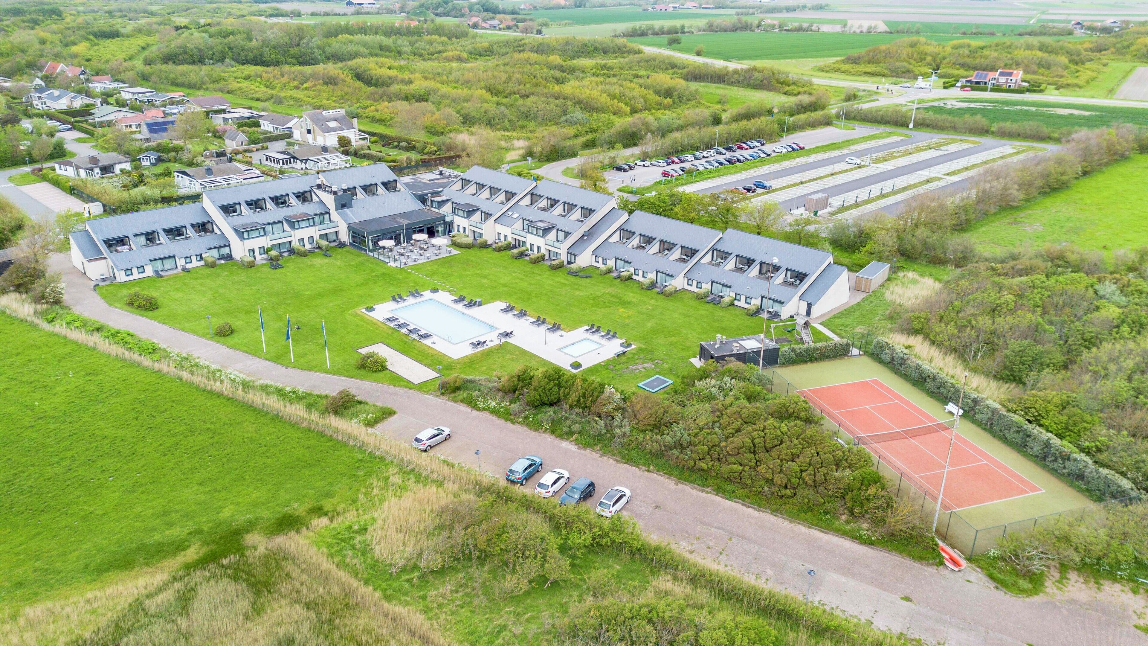 An aerial view of a modern building with multiple sections, including a swimming pool, tennis court, and parking area.