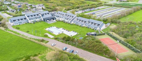An aerial view of a modern building with multiple sections, including a swimming pool, tennis court, and parking area.