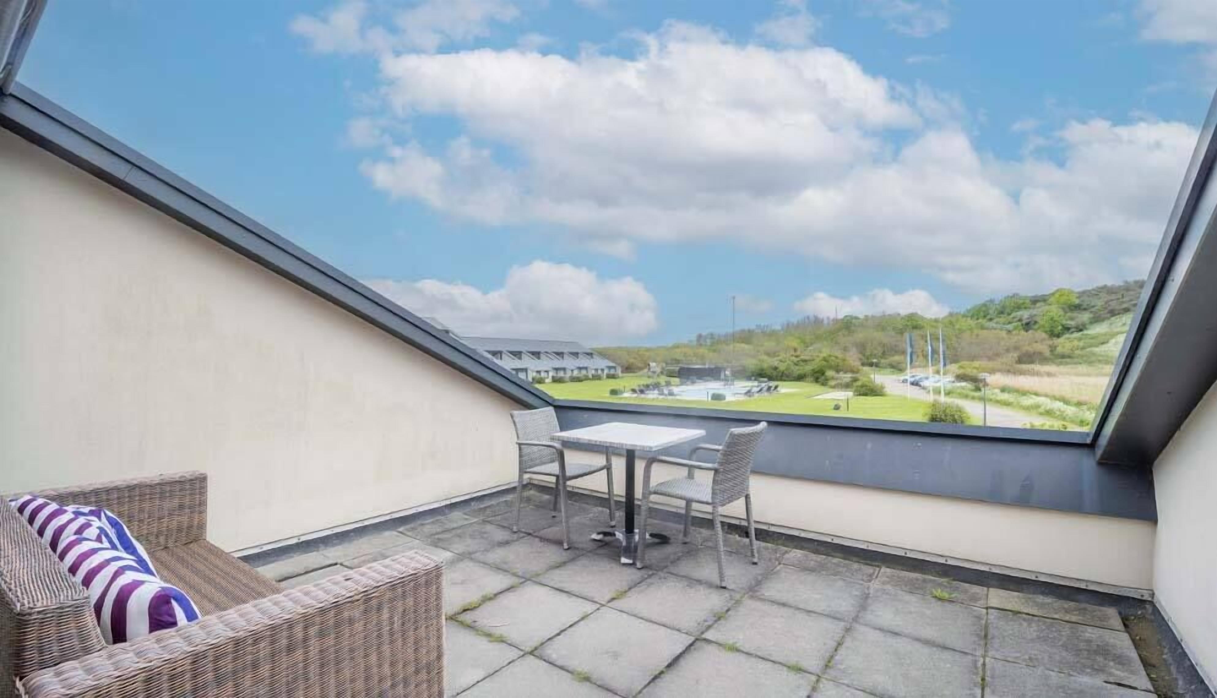 En balkon med en fletsofa, et lille bord og stole, med udsigt over en græsplæne og bygninger.