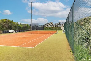 Ein Tennisplatz mit Netz, umgeben von einem grünen Zaun und einem Gebäude mit schrägen Dächern im Hintergrund.
