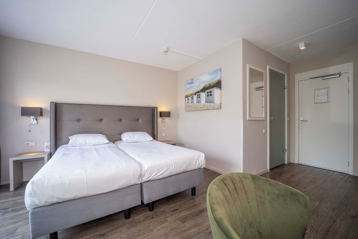 Una cama doble con cabecera gris, dos mesitas de noche con lámparas, una silla verde y un cuadro con casas en la pared.