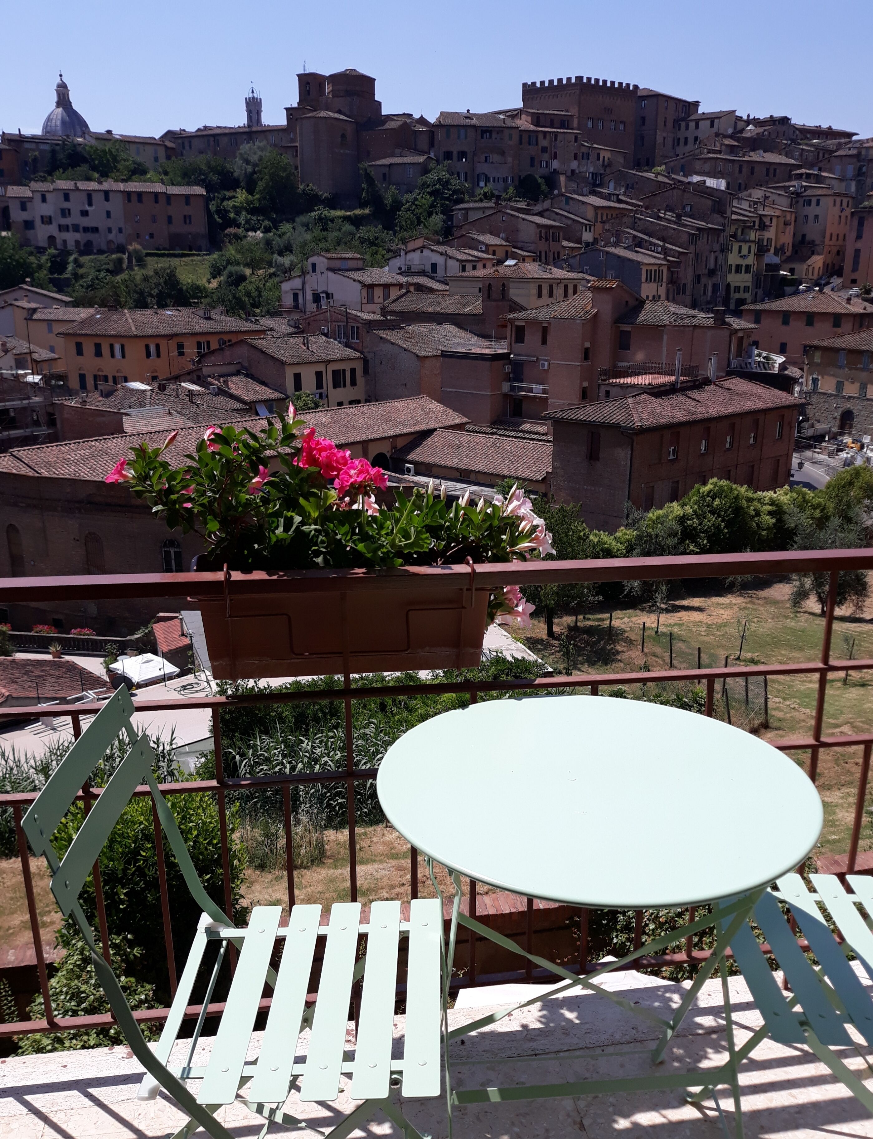 A balcony with a round table and two chairs, overlooking a historic town with terracotta-roofed buildings and a prominent dome structure.