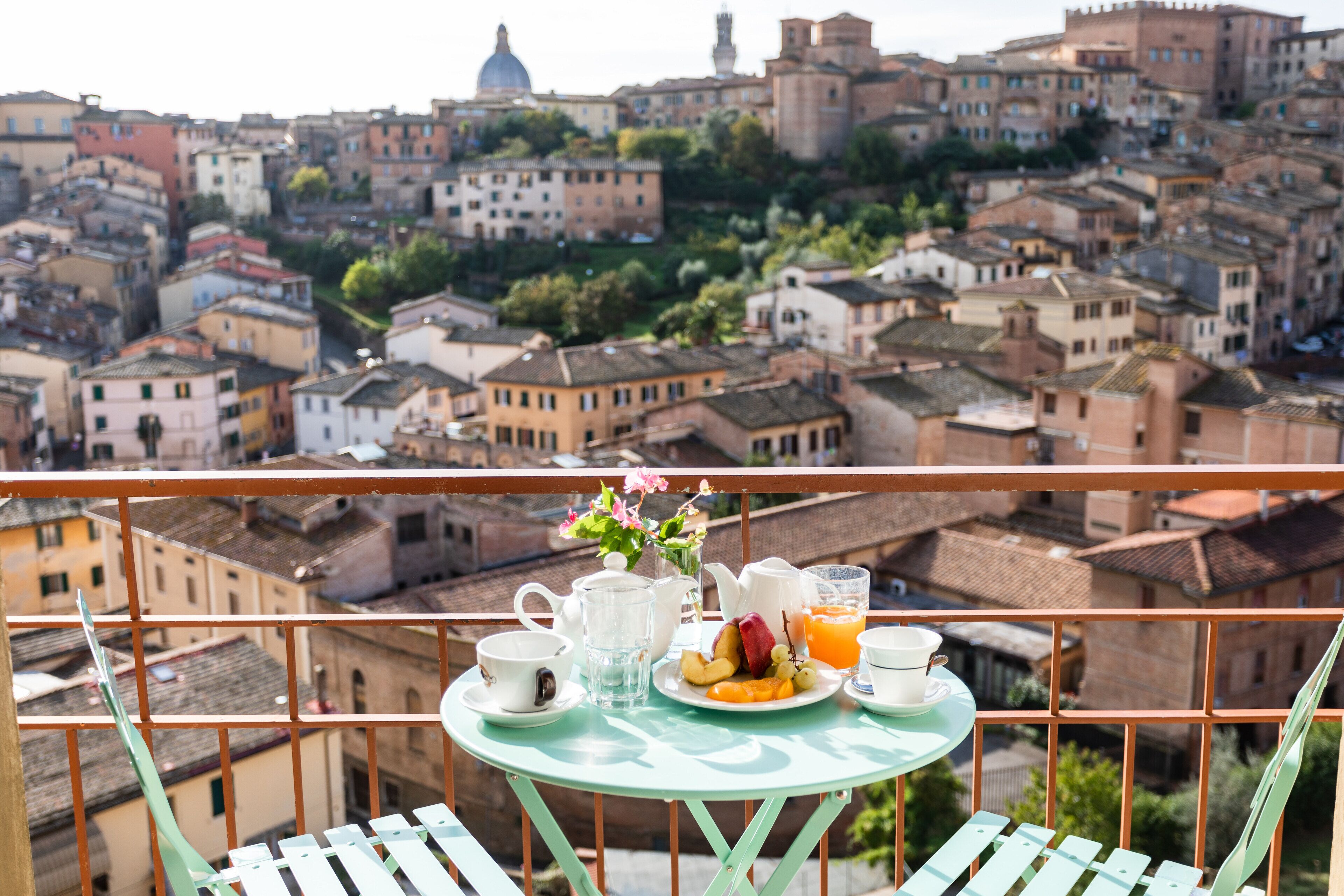 A balcony with a table set for breakfast, overlooking a historic town with terracotta rooftops and a prominent dome structure in the distance.
