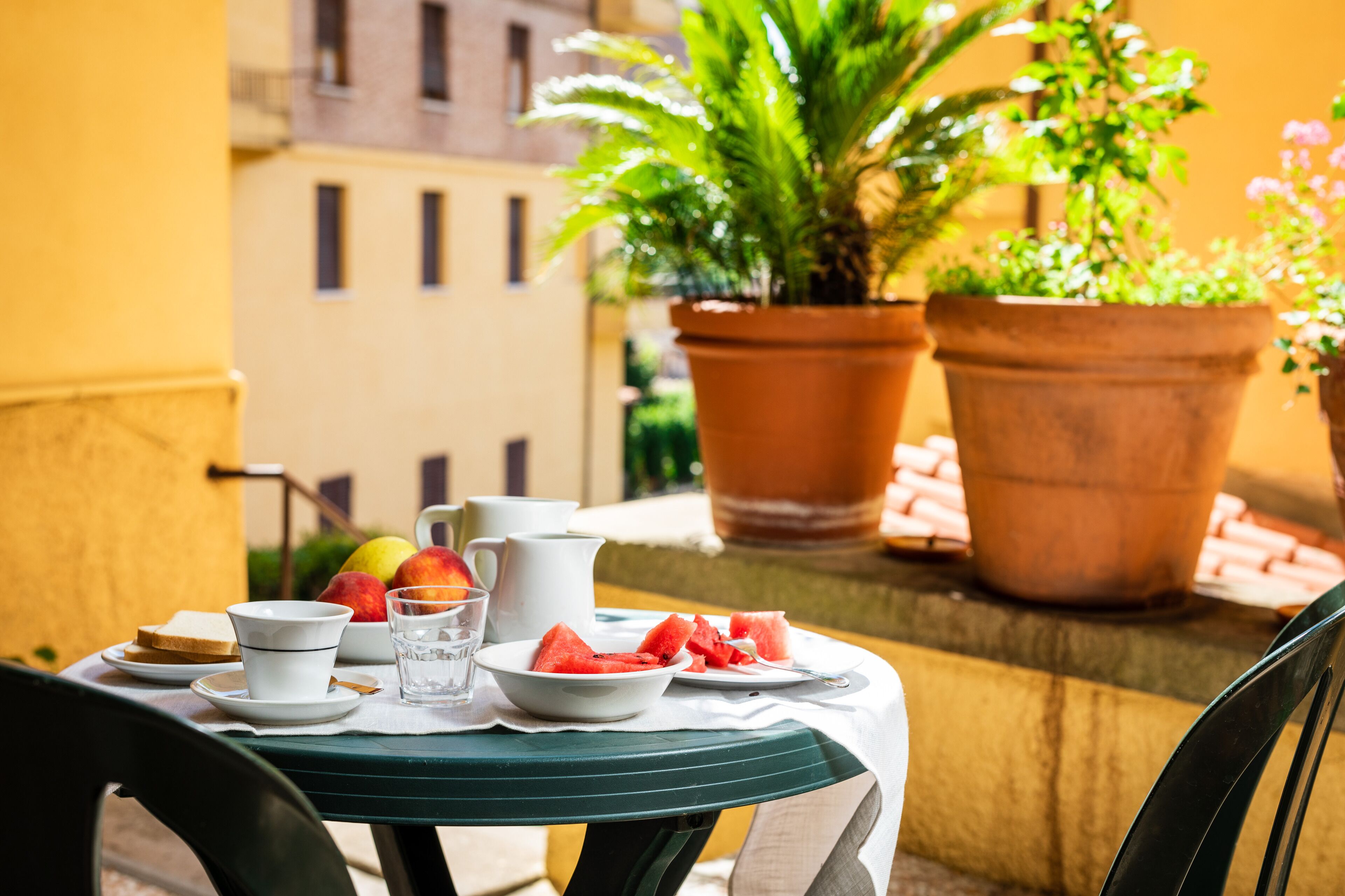 A table set with plates of food, a pitcher, and glasses on a balcony with potted plants and buildings in the background.