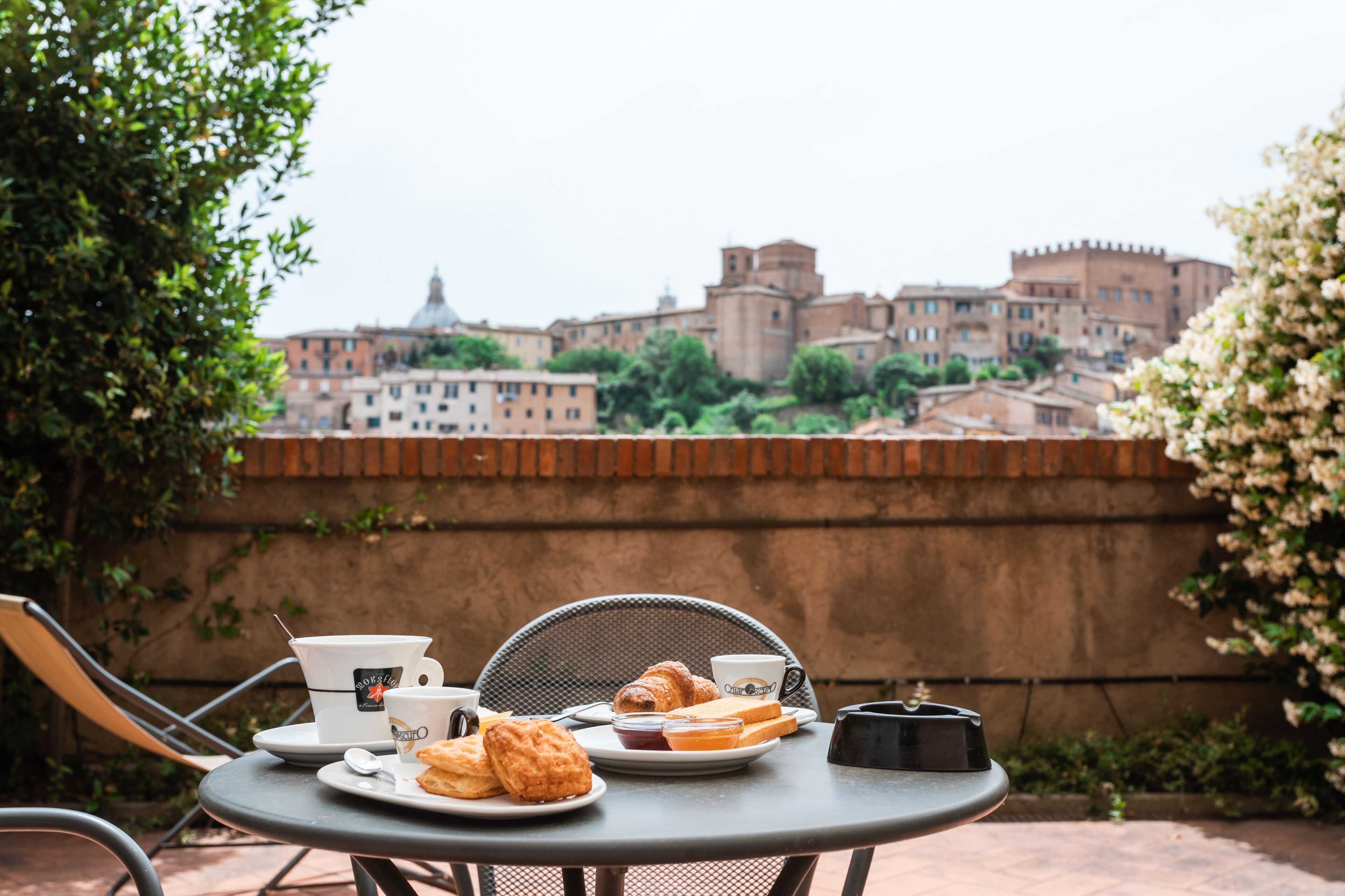 A table with breakfast items and a cup, overlooking a historic cityscape with buildings and a wall.
