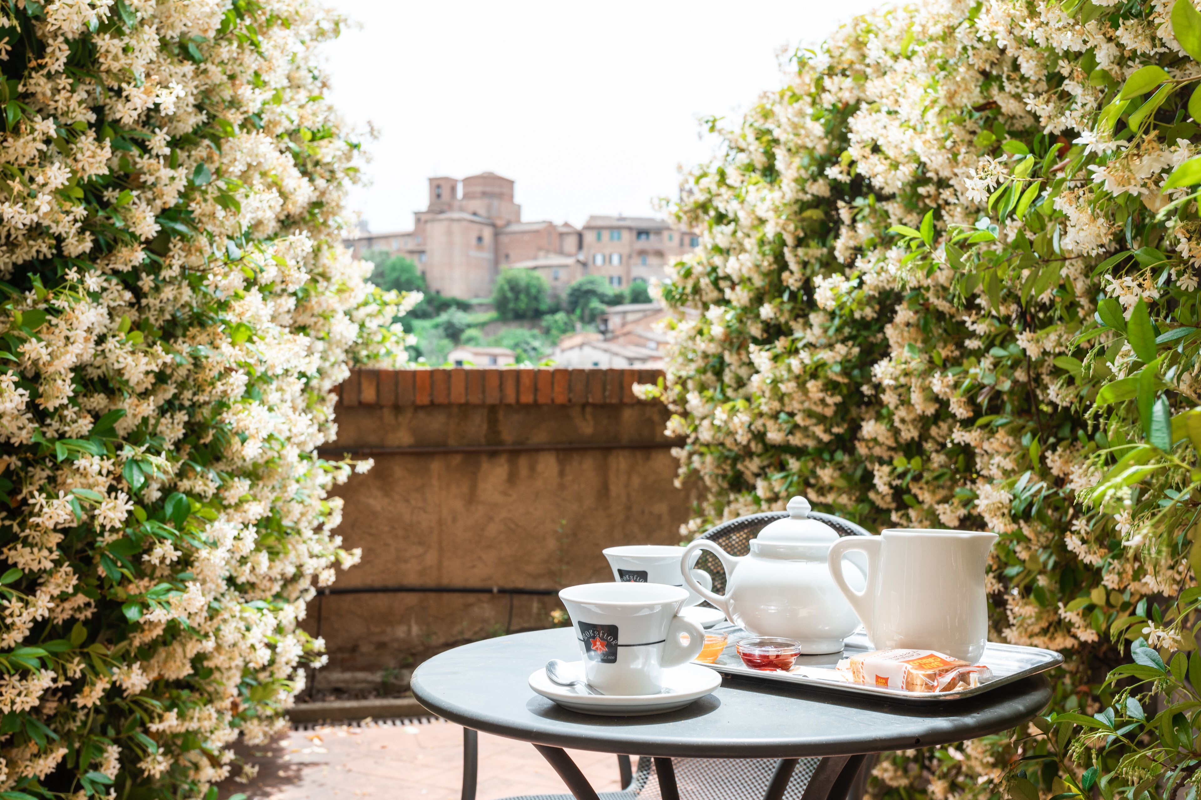 A table set with tea cups, a teapot, and a tray with snacks, surrounded by blooming white flowers, with a view of a historic building in the background.