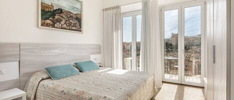 A bedroom with a large bed, a nightstand, a painting on the wall, and a balcony with a view of buildings.